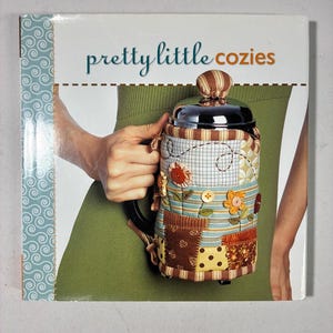 May include: A book titled pretty little cozies featuring a cover image of a quilted fabric tea cosy with a striped top, floral appliques, and button accents wrapped around a metallic French press, set against a light blue patterned border.