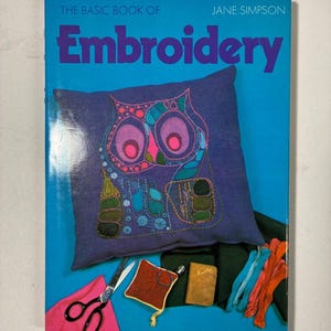 May include: A book titled The Basic Book of Embroidery by Jane Simpson with a bright blue cover. The cover features a purple cushion with a colourful embroidered owl design, metal scissors, a small square pincushion, a needle case, and embroidery thread.