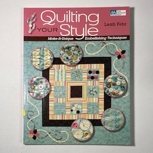 May include: A softcover book titled Quilting Your Style by Leah Fehr. The cover features a light blue quilted background with a central square block pattern, surrounded by six circular insets showing various fabric embellishment techniques.