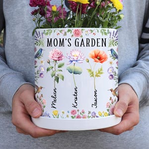 May include: A white ceramic Mom's Garden planter with a floral design, holding colorful flowers. The planter features the names Melvin, Kristen, and Jacon. The planter is held by a person wearing a gray sweatshirt.