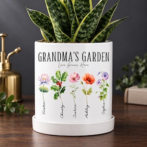 Può includere: Un vaso cilindrico in ceramica bianca con sottovaso abbinato, caratterizzato dalla scritta nera "GRANDMA'S GARDEN" e "Love Grows Here" sopra cinque illustrazioni floreali in stile acquerello, ciascuna etichettata con i nomi Kristen, Christy, Justine, Ashley e Sophia.