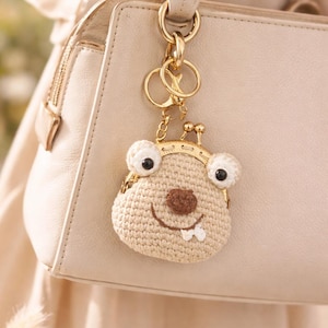 May include: A beige crocheted coin purse with a whimsical face design, featuring large white eyes with black pupils, a brown nose, and a smiling mouth with a single tooth. The purse hangs from a gold-colored keychain attached to a cream-colored handbag.