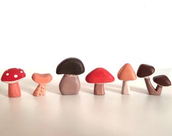 May include: A set of seven hand-painted ceramic mushrooms in various colors and styles. The mushrooms are all different sizes and shapes, and they are arranged in a row on a white background.