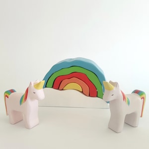 May include: Two white wooden unicorn figurines with rainbow manes and tails stand on a white surface in front of a rainbow-colored wooden arch.