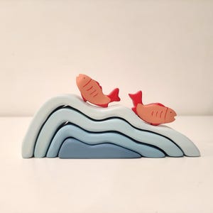 May include: A wooden toy set featuring a blue and white wave stacking toy with two red fish on top. The wave toy is made of five pieces that stack on top of each other.