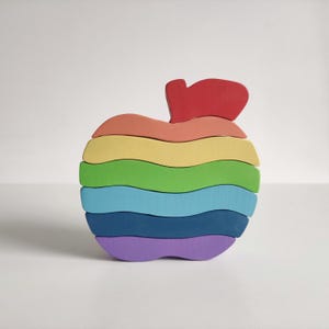 Puede incluir: Un juguete de apilar de madera en forma de manzana arcoíris. La manzana está hecha de siete piezas, cada una de un color diferente del arcoíris. La pieza superior es roja y tiene forma de hoja.