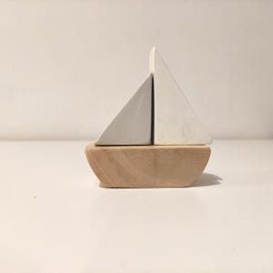 Pode incluir: Um brinquedo de barco à vela de madeira com duas velas triangulares brancas. O barco é feito de madeira natural e tem um casco arredondado.