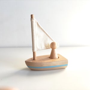 May include: A wooden sailboat toy with a white fabric sail and a small wooden figure standing in the boat. The boat has a blue stripe around the edge.