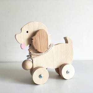 Può includere: Un cane giocattolo di legno con una lingua rosa, orecchie marroni e una corda attaccata a una palla di legno. Il cane ha quattro ruote ed è fatto di legno naturale.