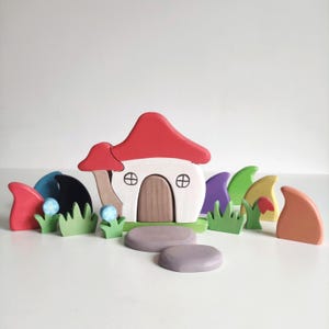May include: A wooden toy set featuring a mushroom house with a red roof, a brown door, and two windows. The set also includes various colored mushrooms, green grass, and two gray stones.
