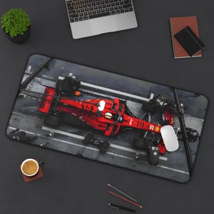 May include: A black and grey mouse mat featuring a red Formula 1 racing car in the centre. The car is surrounded by black and white lines and a grey background. The mouse mat is designed to look like a race track.