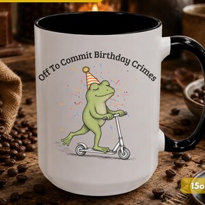 May include: A white ceramic mug with a black interior and handle, featuring a graphic of a green frog wearing a party hat and riding a scooter. The text "Off To Commit Birthday Crimes" is printed above the frog, and the mug is labeled as 15oz.