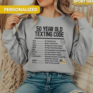 May include: Sport gray sweatshirt with the text "50 Year Old Texting Code" and a list of texting acronyms. The word "Personalized" is printed on a yellow banner. The sweatshirt is worn by a person.