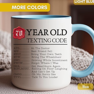May include: A white ceramic mug with a light blue handle and interior, featuring a red wax seal graphic with the number 70 and the text 70 YEAR OLD TEXTING CODE followed by a list of humorous acronym definitions. The mug has an 11oz capacity.