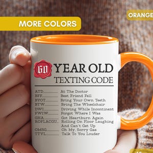 May include: A white 15oz ceramic mug with an orange interior and handle. The front features black text titled 60 YEAR OLD TEXTING CODE, listing humorous acronym definitions like ATD for At The Doctor, BFF for Best Friend Fell, and TTYL for Talk To You Louder.