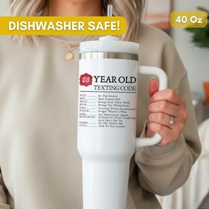 May include: A white 40 oz stainless steel tumbler with a handle and straw. The front features a red wax seal graphic with the text 80 YEAR OLD TEXTING CODE followed by a list of humorous acronym definitions like ATD At The Doctor and TTYL Talk To You Louder.