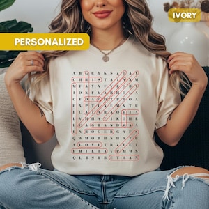May include: Ivory-colored t-shirt with a personalized word search design in red. The word search includes names like "CHRISTA" and "GRANDMA". The shirt has the word "PERSONALIZED" printed on it.