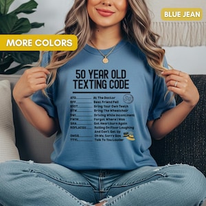 May include: A light blue t-shirt with the text "50 YEAR OLD TEXTING CODE" and a list of texting acronyms. The shirt also has the words "MORE COLORS" and "BLUE JEAN" in the top corners.