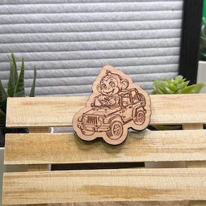 May include: A wooden monkey driving a Jeep cutout. The monkey is smiling and the Jeep has a spare tyre on the back. The cutout is light brown and sits on a wooden pallet.