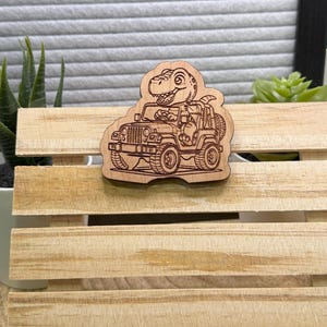 May include: Wooden laser-cut dinosaur driving a Jeep. The dinosaur has a friendly expression. The Jeep has detailed wheels and a spare tyre. The wooden cutout is placed on a wooden crate.