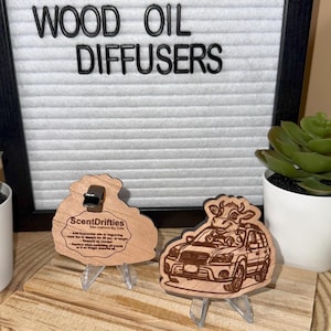 Pode incluir: Difusores de óleo de madeira com o texto "WOOD OIL DIFFUSERS" em letras pretas. São exibidos dois ambientadores de carro de madeira, um com uma vaca dirigindo um carro. O outro tem o texto "ScentDrifties".