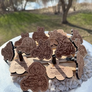 May include: Wooden car air fresheners with dog breeds driving jeeps. Each air freshener is laser-cut with detailed designs and includes the text "ScentDrifties". The air fresheners are displayed on a wooden stand with white stones.