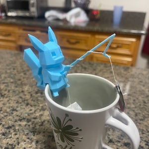May include: A light blue 3D printed Pokemon character holding a fishing rod over a white mug with a green flower design and the word "coffee" printed on it. The mug has a tea bag inside.