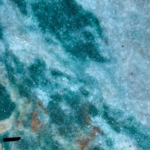 May include: Close-up of a polished stone with a swirling pattern of teal and white. The stone has a natural, textured appearance with areas of darker teal and hints of brown. The overall effect is reminiscent of flowing water or a marbled surface. Ideal for jewelry or decorative purposes.