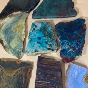 May include: Assortment of raw, polished gemstones in various shapes and colors. Includes pieces in shades of blue, green, brown, and red. The stones have unique patterns and textures, showcasing natural mineral formations.