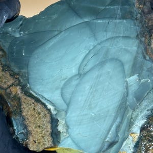 May include: A large, irregularly shaped stone with a smooth, light blue surface. The stone has a layered appearance with darker blue and brown sections along the edges. The stone's surface has a polished look, with natural patterns and textures.