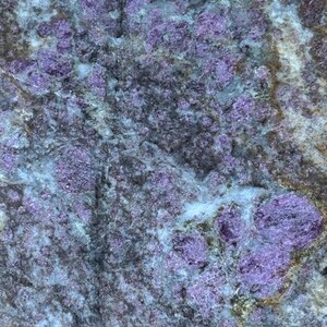 May include: Close-up of a natural stone with a mix of colors. The stone features a blend of purple, gray, and light blue hues. The surface has a textured appearance with various patterns and formations.