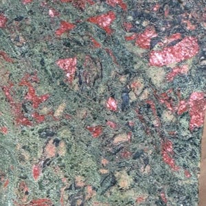 May include: A polished slab of eclogite rock featuring a dense, mottled texture. The surface displays a complex mixture of vibrant red garnet crystals embedded within a dark, forest green matrix of omphacite, with small black mineral inclusions.