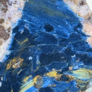May include: A polished slab of blue pietersite gemstone featuring deep navy and royal blue swirling patterns with streaks of golden yellow and brown mineral inclusions. The surface displays a chatoyant, fibrous texture with a glossy finish.