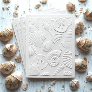 May include: A stack of white embossed cards featuring various seashell designs, including conch shells, scallops, and sand dollars. The cards are surrounded by real seashells on a light blue wooden surface, creating a coastal theme.