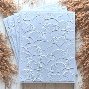May include: A stack of light blue embossed paper sheets featuring a repeating ginkgo leaf pattern. The leaves have detailed veining and a raised texture. The paper is stacked on a light wood surface with dried plants in the background.
