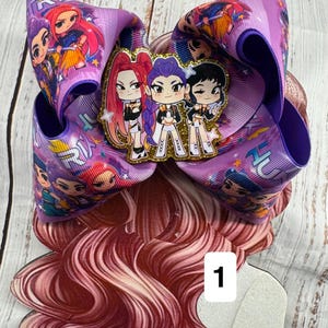 May include: A large purple hair bow featuring a repeating pattern of cartoon characters with red, purple, and black hair. A central glittery acrylic charm displays three stylized cartoon figures in matching outfits with the number 1 on a white square.