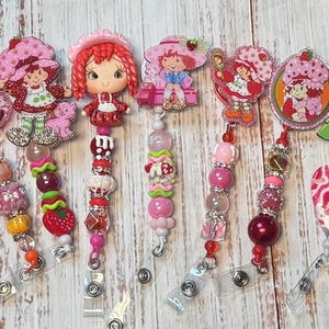 Puede incluir: Una colección de portatarjetas retráctiles con el tema de Strawberry Shortcake. Cada soporte presenta un diseño diferente del personaje, con varios acentos de cuentas en tonos de rosa, rojo y blanco. Los carretes de la tarjeta de identificación están sujetos a un clip plateado.