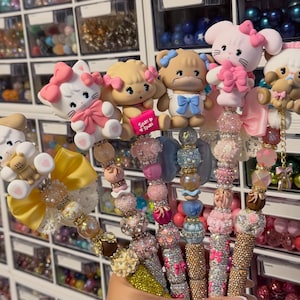 May include: A selection of decorative pens with cartoon animal toppers, such as a cat, dog, and bear. The pens have beaded shafts in various colours, with bows and other embellishments. The background shows storage containers filled with colourful beads.