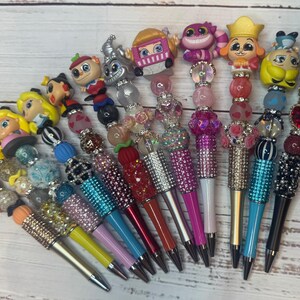 May include: A collection of decorative pens with character-themed toppers. The pens feature colorful beads and rhinestone accents. The pen barrels are various colors, including pink, blue, and yellow. The toppers depict cartoon characters.