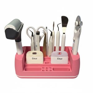 May include: A pink Cricut tool organiser filled with various crafting tools. The tools include scissors, tweezers, a scraper, a rotary cutter, and a roller. The organiser has two compartments with the Cricut logo.