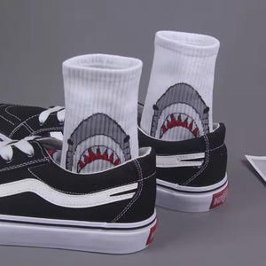 May include: A pair of white socks with a shark graphic on each sock. The socks are worn with black sneakers.