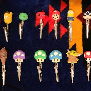 May include: A collection of novelty clips featuring various cartoon characters and food items. The clips include a burger, ice cream cone, mushroom, and characters like Mario, Baby Yoda, and Minion. The clips are silver and are arranged on a patterned surface.