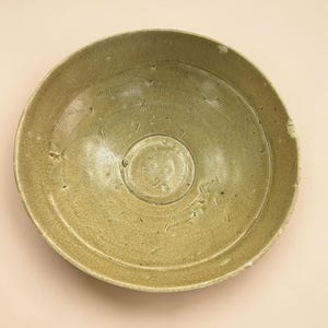 May include: A round ceramic bowl with a muted olive-green glaze and a visible crackle finish. The interior surface features a series of impressed, repeating comb-like patterns arranged in a circular motion around a smooth, plain central medallion.