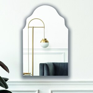 May include: A wall-mounted mirror featuring a scalloped arch top design. The reflective surface displays a room interior with a gold-toned floor lamp and a dark-colored sofa. The mirror has a clean, frameless edge and a vertical orientation.
