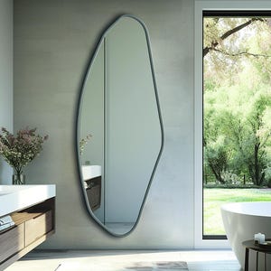 May include: A full-length wall mirror featuring an irregular, organic, pebble-like shape with a thin, dark gray frame. The reflective glass surface displays a clear, undistorted view of the surrounding interior space, mounted vertically on a wall.