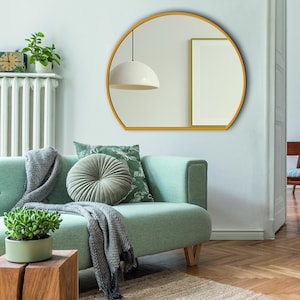 Peut inclure: Un miroir mural semi-circulaire doté d'un cadre fin en métal doré. Le miroir présente un bord inférieur plat et un sommet incurvé, reflétant une suspension blanche et un cadre photo sur le mur dans sa surface en verre.