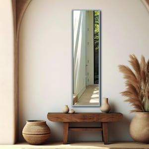 May include: A full-length mirror with a light gray frame, standing on a wooden bench with two vases and a woven basket. The mirror reflects a hallway with a white door and a window with sunlight streaming through.
