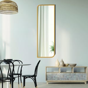 May include: A tall, rectangular mirror with a rounded top and a gold frame. The mirror is hanging on a white wall above a wooden cabinet with a tiled front.