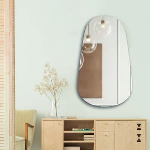 May include: A wall-mounted mirror with a rounded, elongated shape. The reflective surface shows a room interior with two hanging glass globe pendant lights and a brown rectangular panel. The mirror has a smooth, frameless edge design.