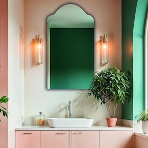 May include: A wall-mounted mirror with a rounded arch top, featuring a reflective glass surface. The mirror is positioned above a white rectangular sink basin and a light pink vanity cabinet, flanked by two vertical cylindrical wall sconces.